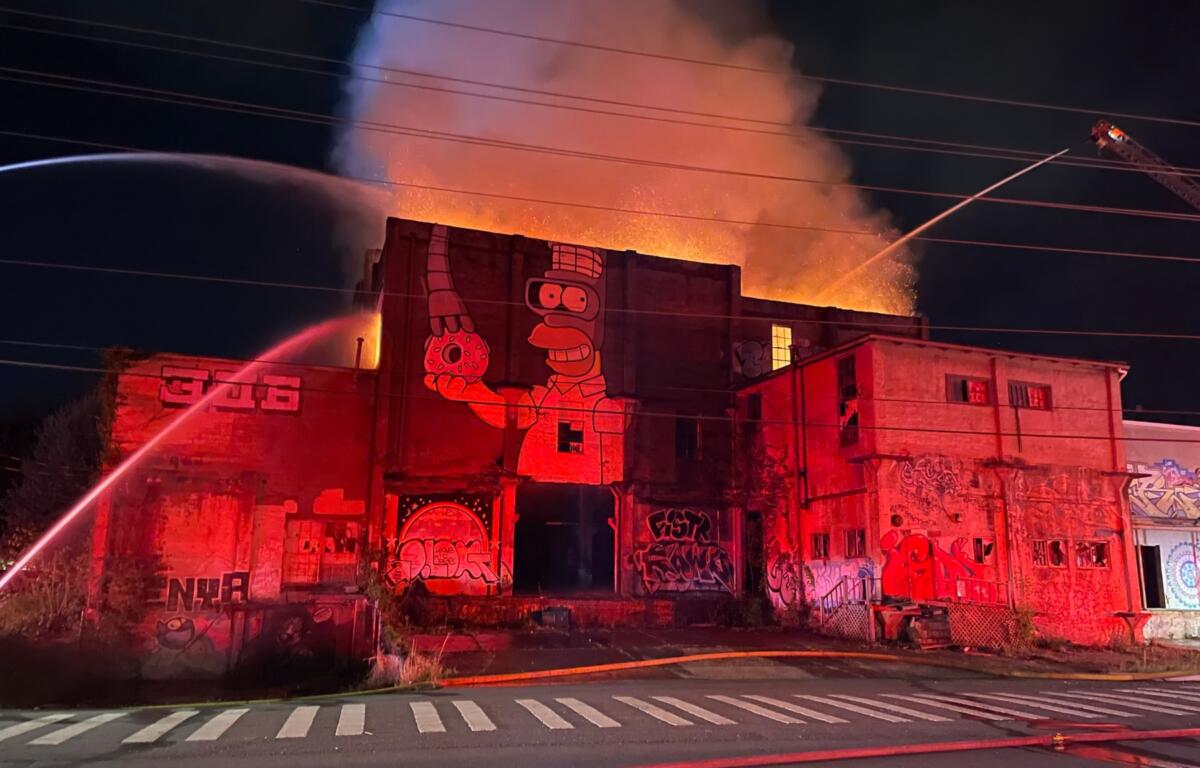 Nighttime fire engulfs a graffiti-covered building; flames and smoke erupt from the roof as firefighters spray water from hoses.