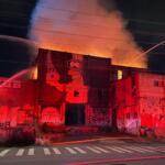 Nighttime fire engulfs a graffiti-covered building; flames and smoke erupt from the roof as firefighters spray water from hoses.
