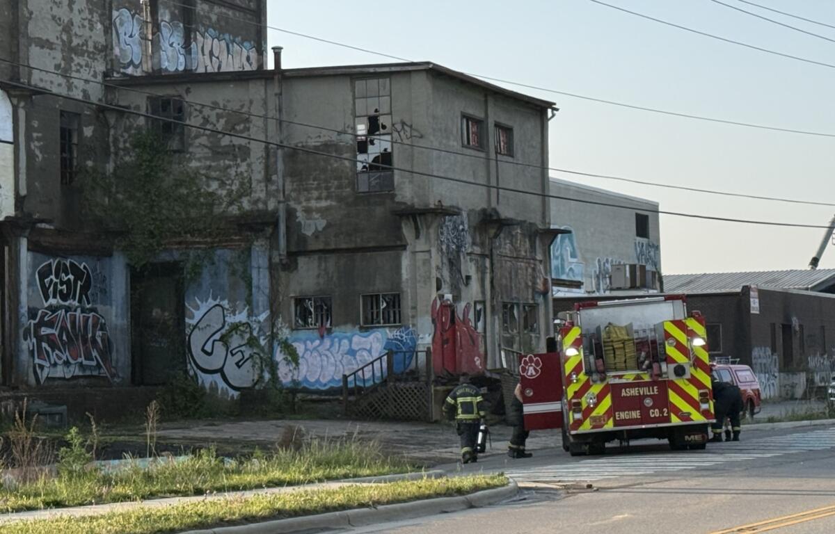 Firefighters in turnout gear near a graffiti-covered, dilapidated two-story building with a red fire engine on the street.