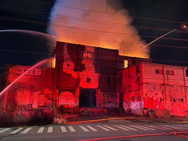 Nighttime fire engulfs a graffiti-covered building; flames and smoke erupt from the roof as firefighters spray water from hoses.