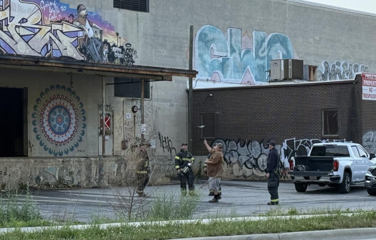 Firefighters stand in an urban alley beside graffiti-covered walls and a brick building, with a white pickup parked nearby.