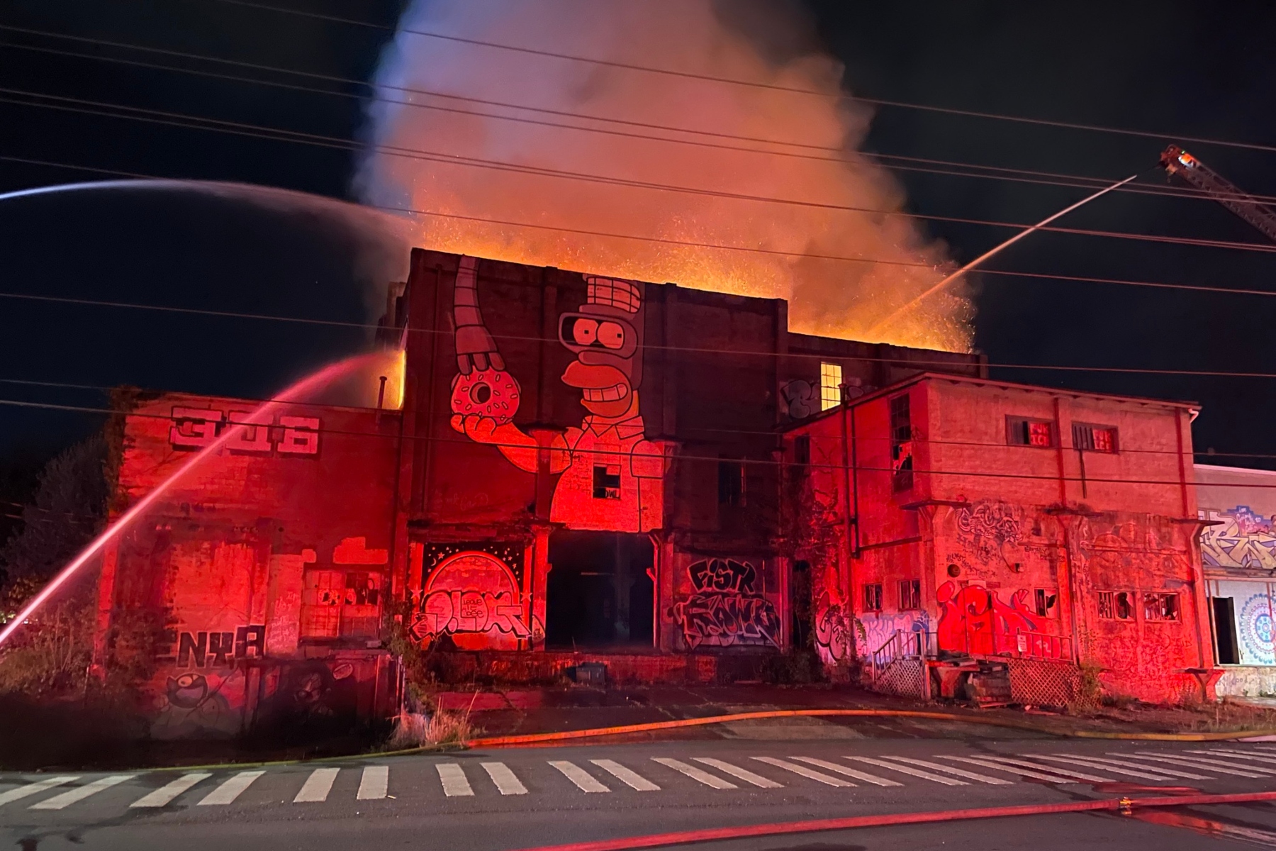 Nighttime fire engulfs a graffiti-covered building; flames and smoke erupt from the roof as firefighters spray water from hoses.