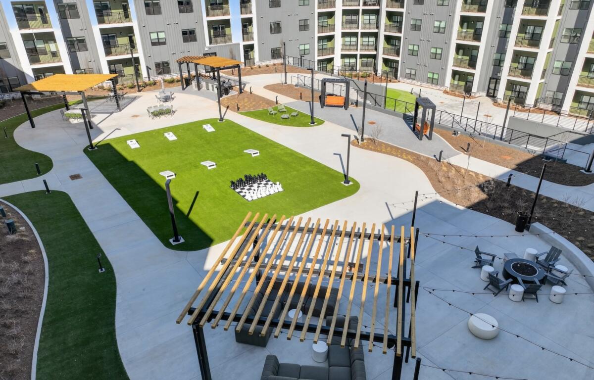 Modern apartment courtyard with green lawn, concrete paths, and a large chessboard set of pieces in the center surrounded by seating areas and pergolas.