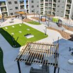 Modern apartment courtyard with green lawn, concrete paths, and a large chessboard set of pieces in the center surrounded by seating areas and pergolas.