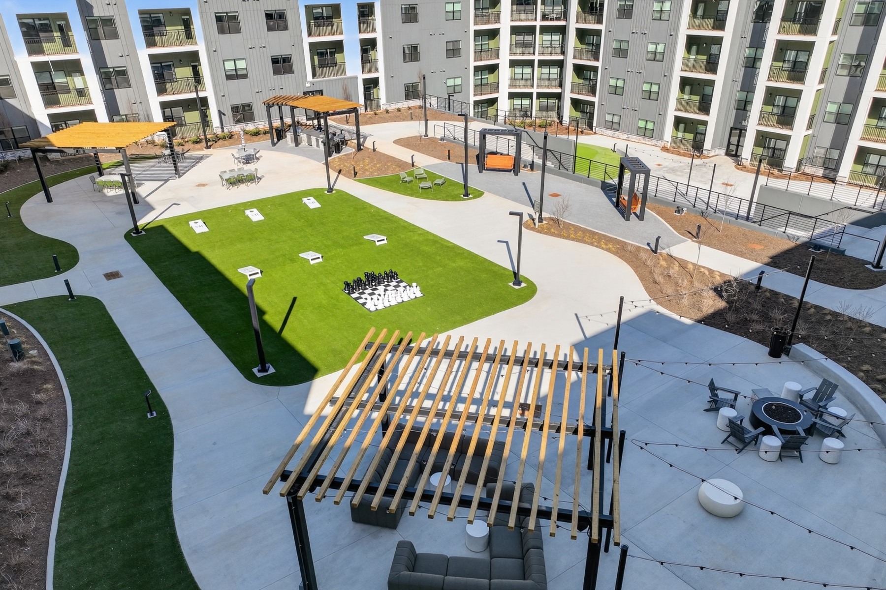 Modern apartment courtyard with green lawn, concrete paths, and a large chessboard set of pieces in the center surrounded by seating areas and pergolas.