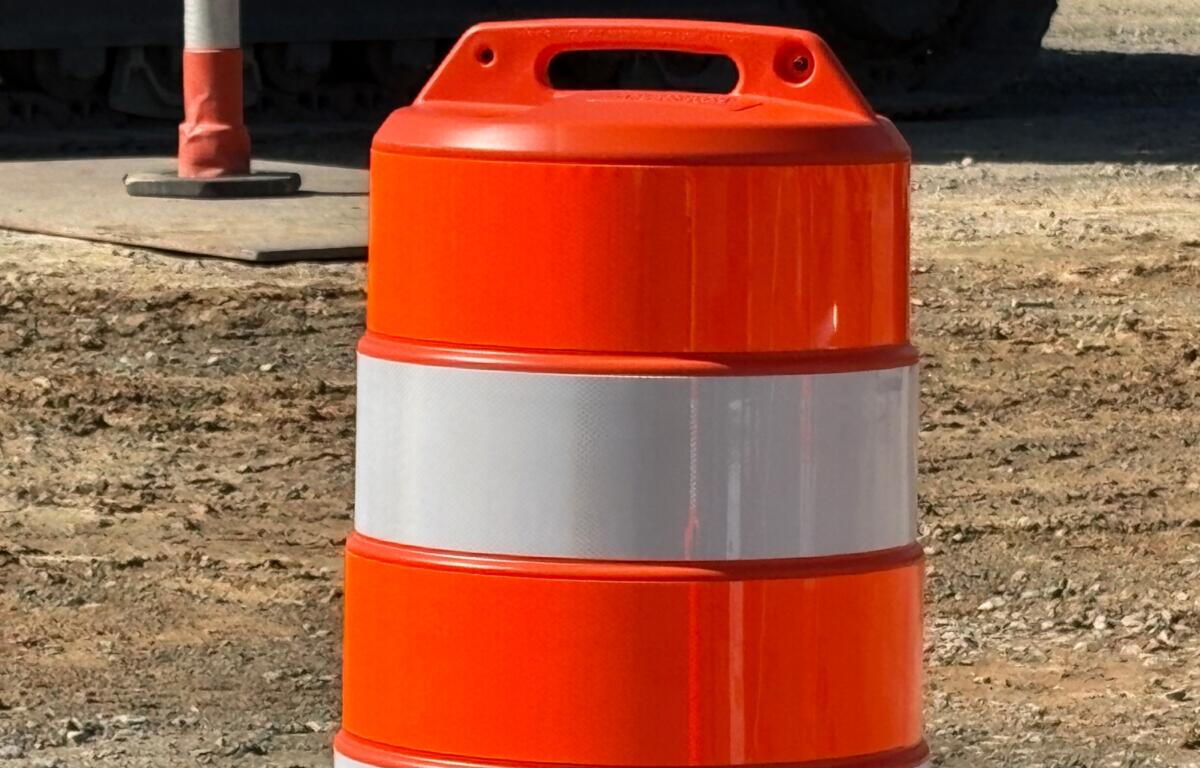 Orange construction barrel with a reflective white band, placed on a dirt site with equipment in the background