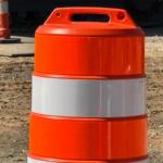 Orange construction barrel with a reflective white band, placed on a dirt site with equipment in the background