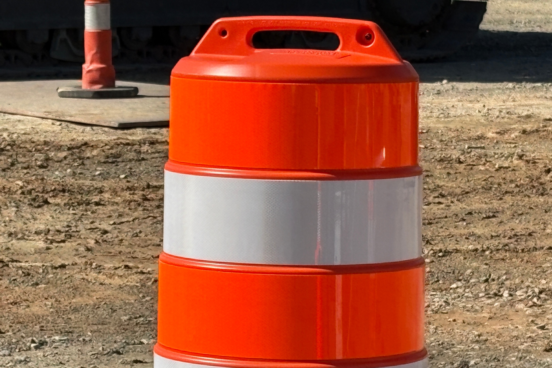 Orange construction barrel with a reflective white band, placed on a dirt site with equipment in the background