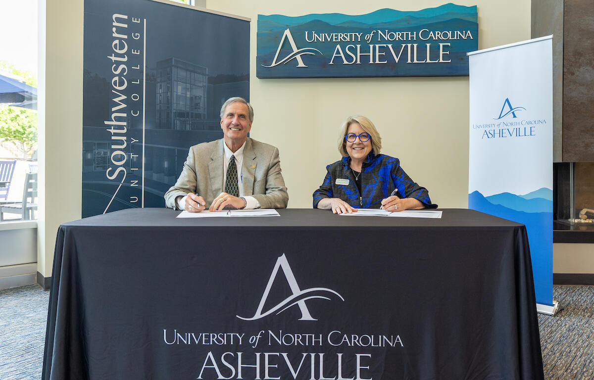 Two professionals sign documents at a head table for a UNC Asheville event, with banners in the background.