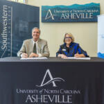 Two professionals sign documents at a head table for a UNC Asheville event, with banners in the background.