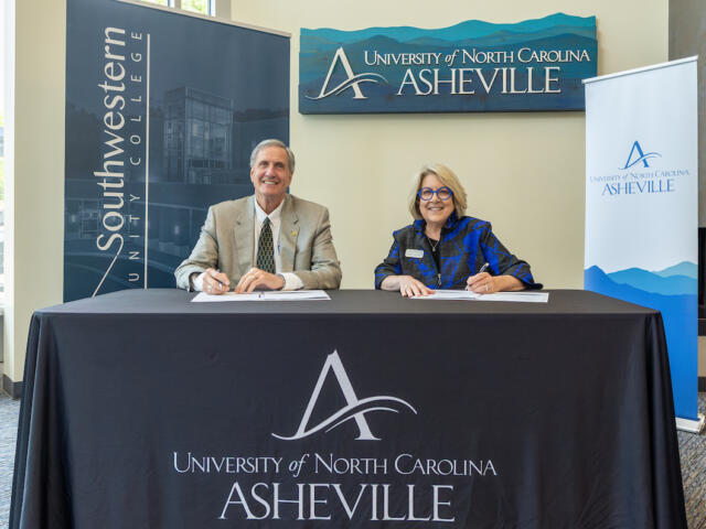 Two professionals sign documents at a head table for a UNC Asheville event, with banners in the background.
