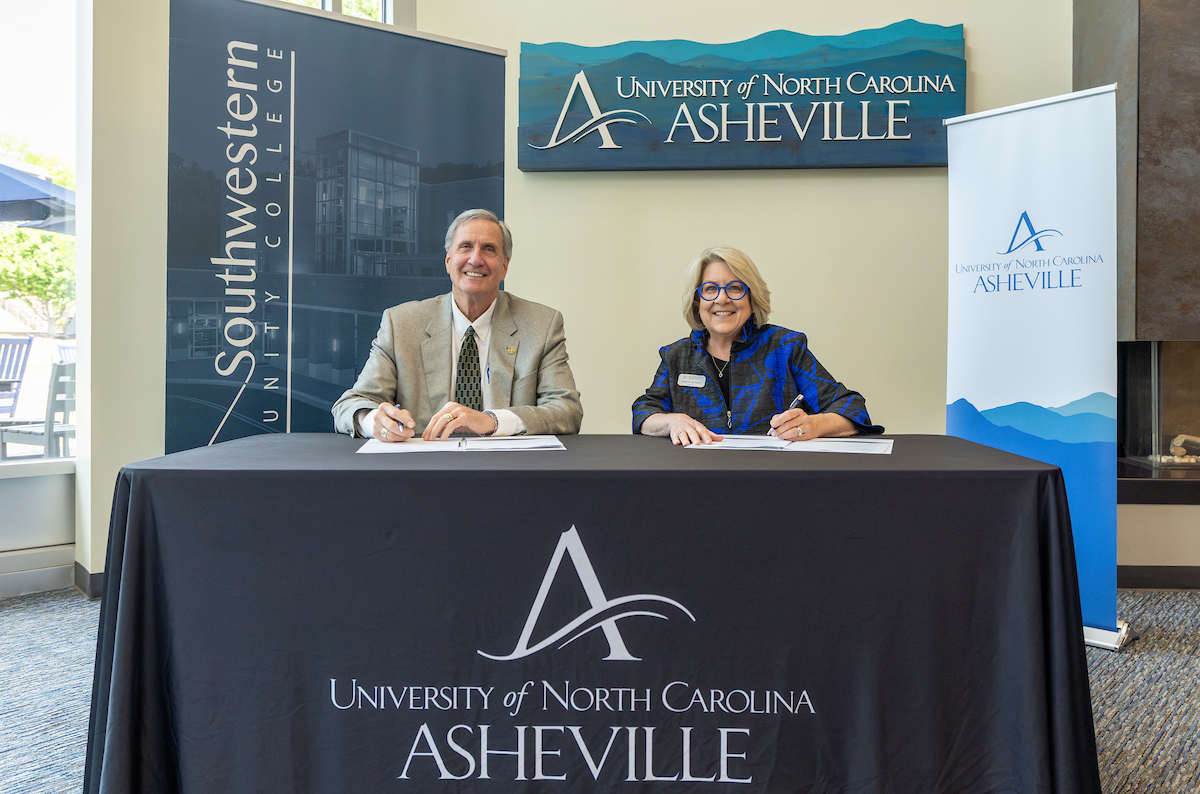 Two professionals sign documents at a head table for a UNC Asheville event, with banners in the background.