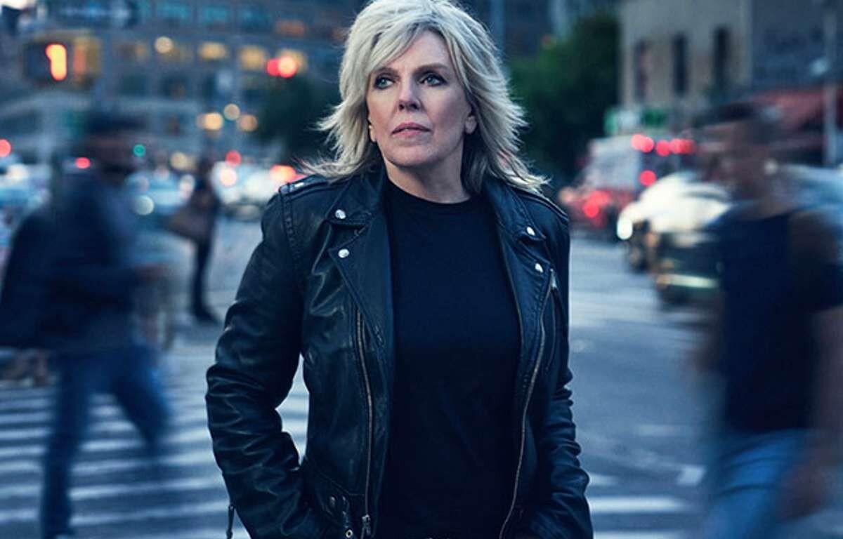 Woman in a black leather jacket stands in a busy city street, looking ahead as blurred pedestrians pass by.