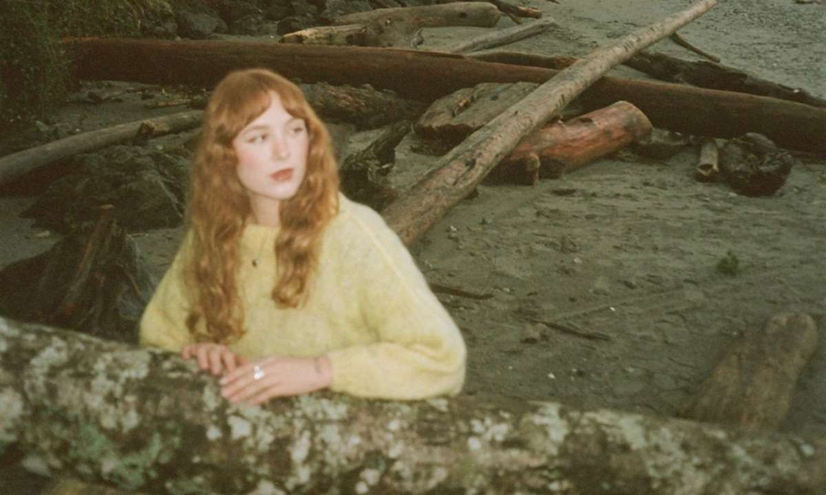 Woman with long red hair in a pale yellow sweater leans on a mossy log at a driftwood-strewn beach socal scene.