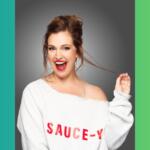 Cheerful woman with red lipstick wearing a white off-shoulder sweater that reads 'SAUCE-Y' against a gray studio backdrop, laughing and pulling a strand of hair.