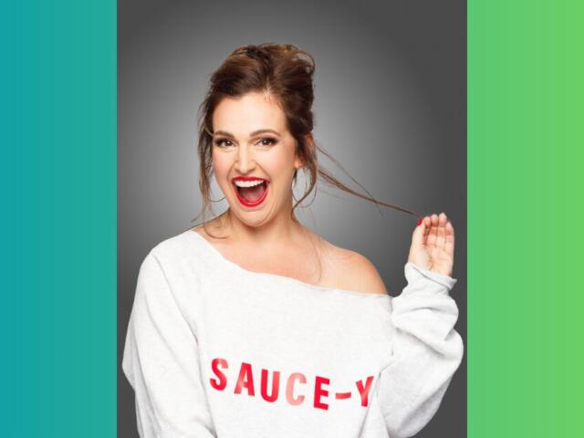 Cheerful woman with red lipstick wearing a white off-shoulder sweater that reads 'SAUCE-Y' against a gray studio backdrop, laughing and pulling a strand of hair.