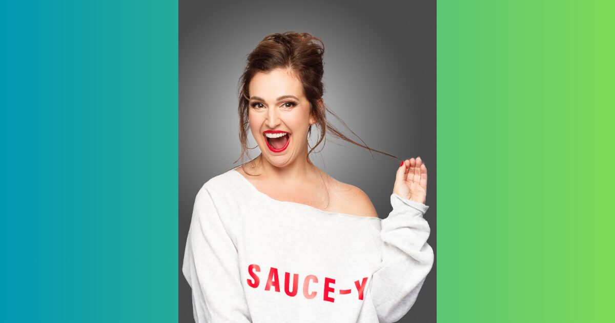 Cheerful woman with red lipstick wearing a white off-shoulder sweater that reads 'SAUCE-Y' against a gray studio backdrop, laughing and pulling a strand of hair.