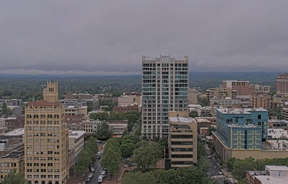 Downtown skyline with a tall glass high-rise in the center, surrounded by shorter brick and beige buildings under a cloudy sky.