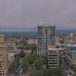Downtown skyline with a tall glass high-rise in the center, surrounded by shorter brick and beige buildings under a cloudy sky.