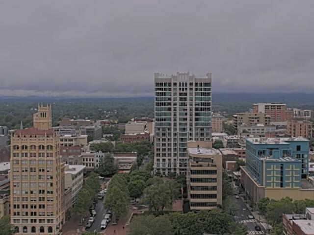 Downtown skyline with a tall glass high-rise in the center, surrounded by shorter brick and beige buildings under a cloudy sky.