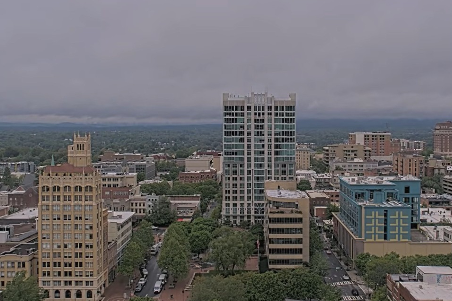 Downtown skyline with a tall glass high-rise in the center, surrounded by shorter brick and beige buildings under a cloudy sky.