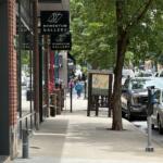 Urban sidewalk with Momentum Gallery signs, brick storefronts, and trees; pedestrians walk along the sunny street.