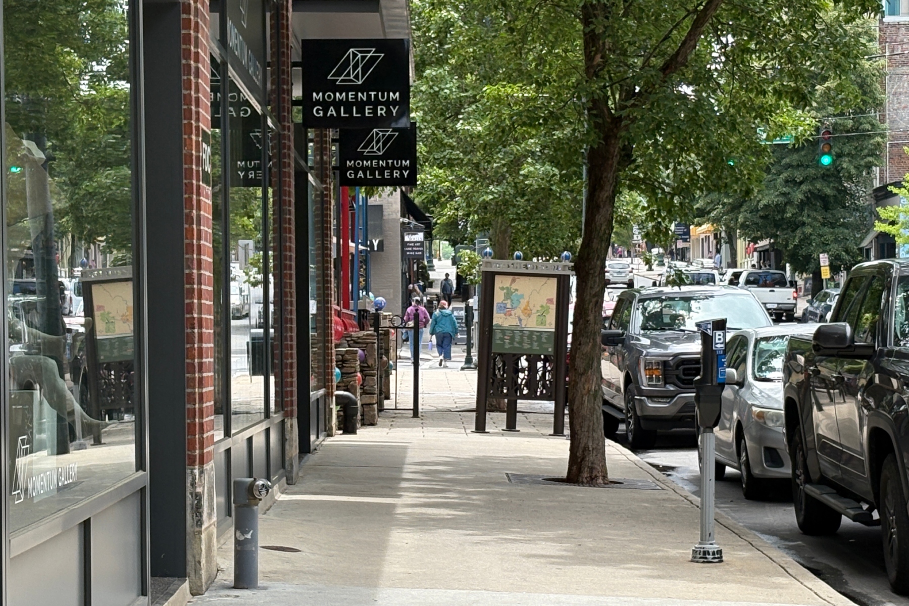 Urban sidewalk with Momentum Gallery signs, brick storefronts, and trees; pedestrians walk along the sunny street.