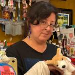 Woman with glasses holding a white plush fox in a colorful gift shop filled with stickers and candy.