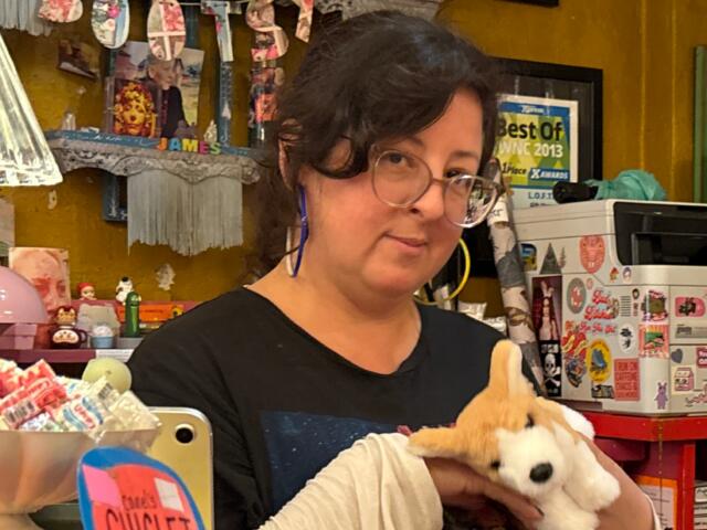 Woman with glasses holding a white plush fox in a colorful gift shop filled with stickers and candy.