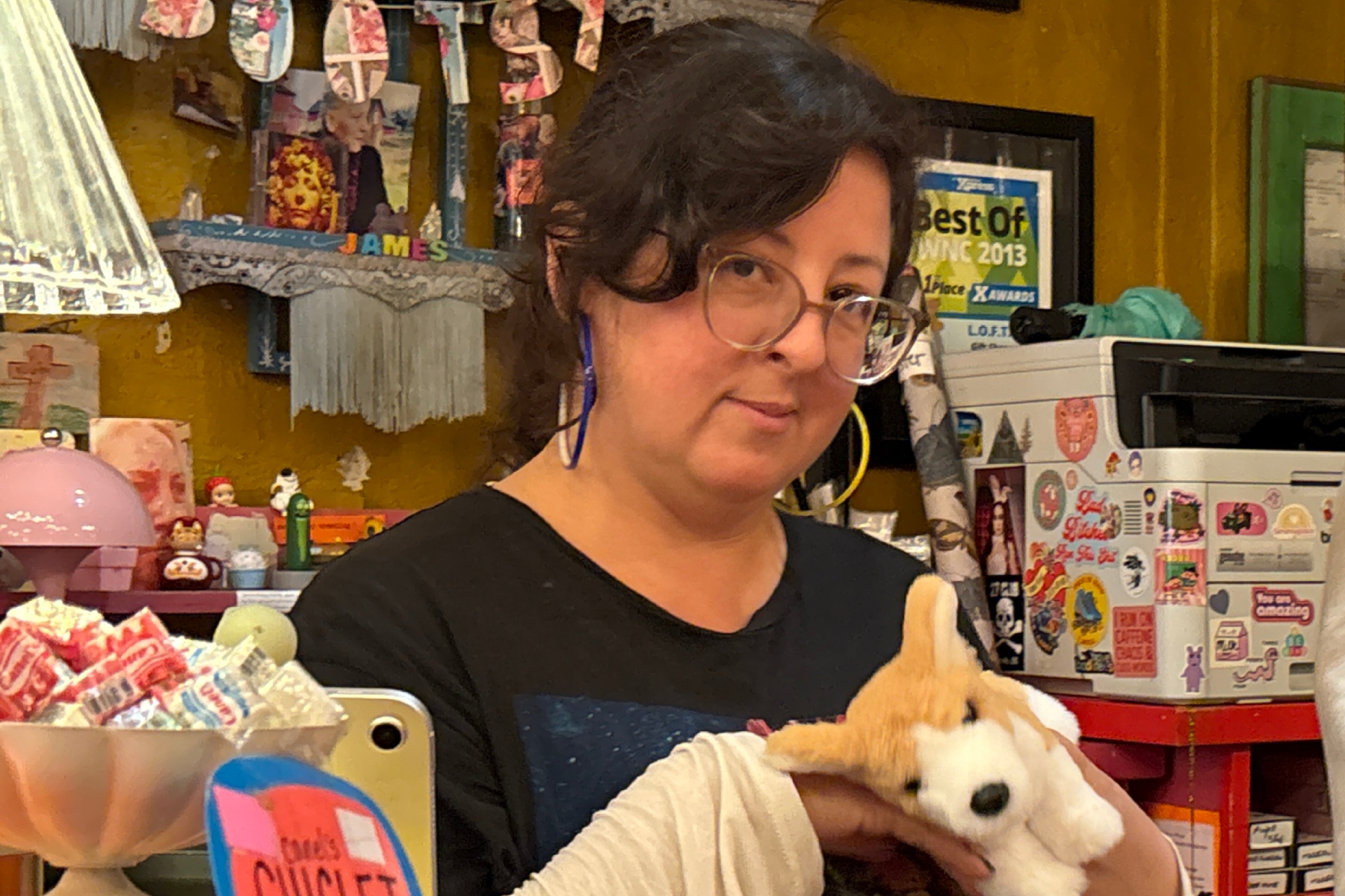 Woman with glasses holding a white plush fox in a colorful gift shop filled with stickers and candy.