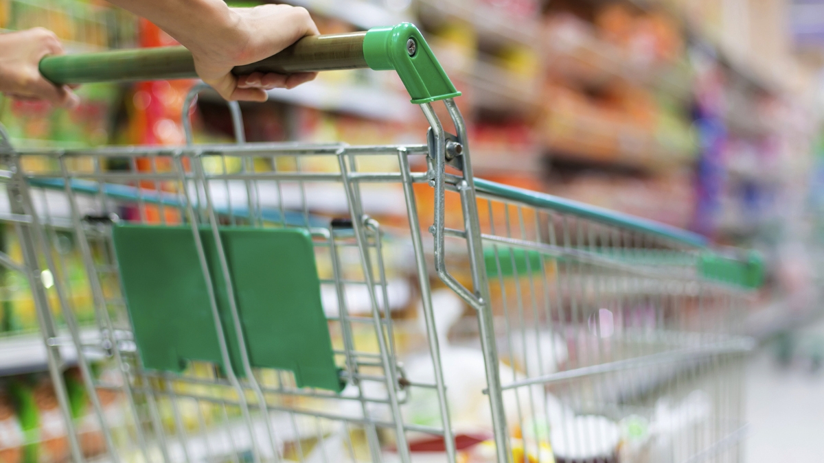 Shoppers push a shopping cart down a grocery store aisle with blurred shelves in the background.