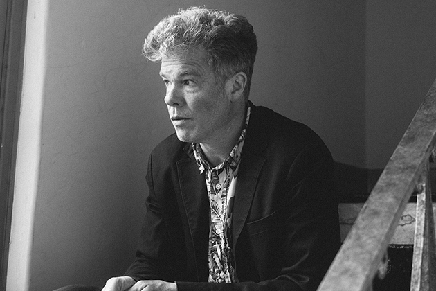 Black-and-white photo of a man in a blazer sitting near a stair railing, looking to the left.