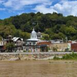 Riverside town with brick storefronts along a stone embankment, trees on a hill behind, under a blue sky with scattered clouds.