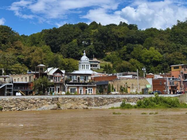 Riverside town with brick storefronts along a stone embankment, trees on a hill behind, under a blue sky with scattered clouds.