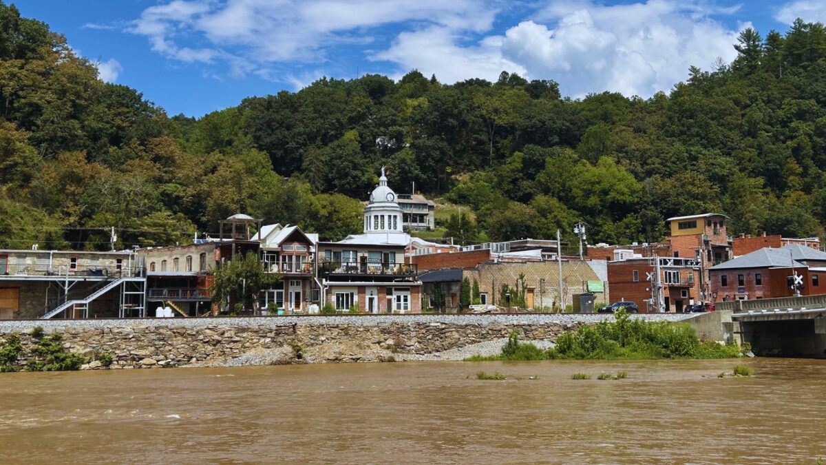 Riverside town with brick storefronts along a stone embankment, trees on a hill behind, under a blue sky with scattered clouds.