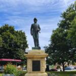 Bronze statue of a male military figure in uniform atop a stone pedestal in a sunny park, with trees and a small pavilion nearby.