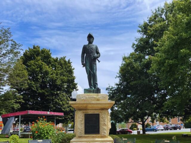 Bronze statue of a male military figure in uniform atop a stone pedestal in a sunny park, with trees and a small pavilion nearby.