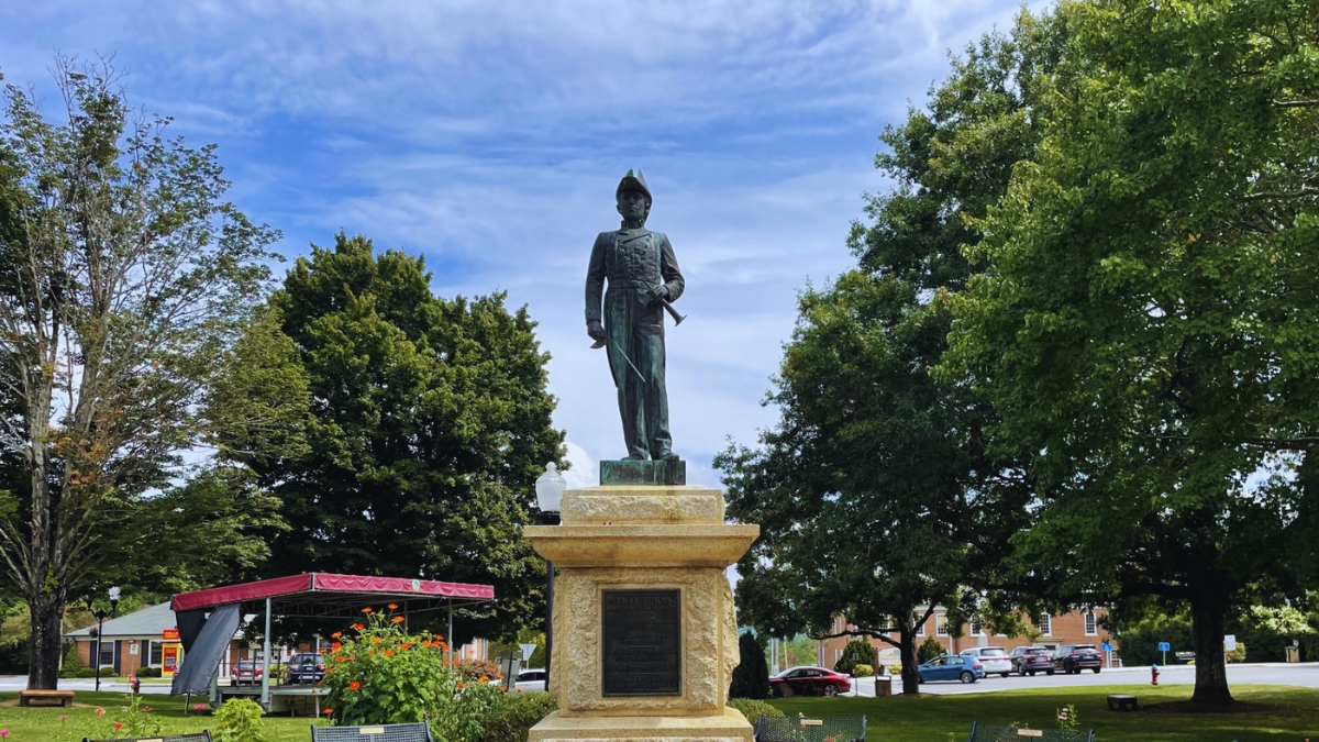 Bronze statue of a male military figure in uniform atop a stone pedestal in a sunny park, with trees and a small pavilion nearby.