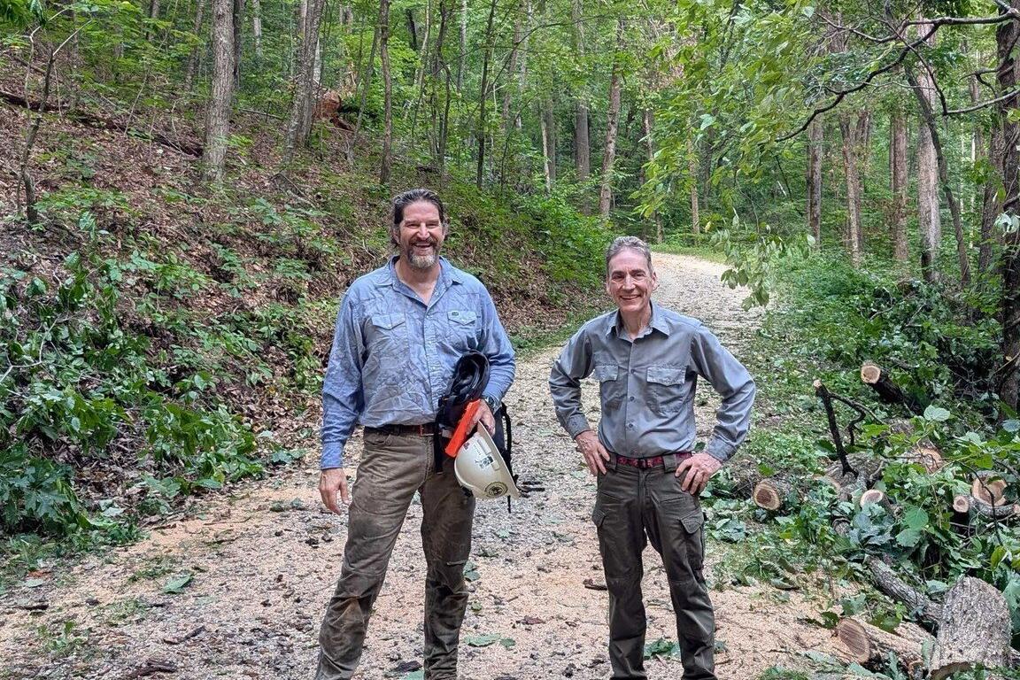 Two men in work shirts stand on a forest gravel path, smiling, with cut branches and tools nearby on the right side.