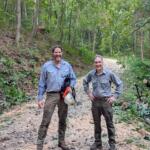 Two men in work shirts stand on a forest gravel path, smiling, with cut branches and tools nearby on the right side.