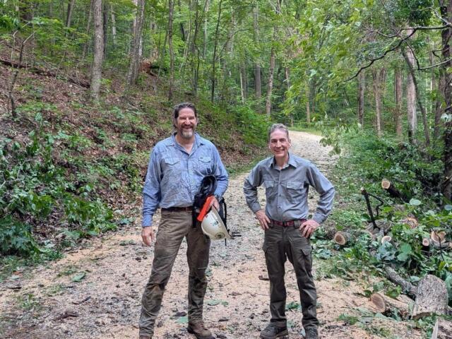 Two men in work shirts stand on a forest gravel path, smiling, with cut branches and tools nearby on the right side.