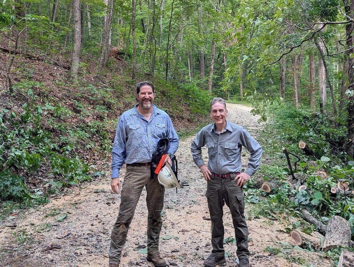 Two men in work shirts stand on a forest gravel path, smiling, with cut branches and tools nearby on the right side.