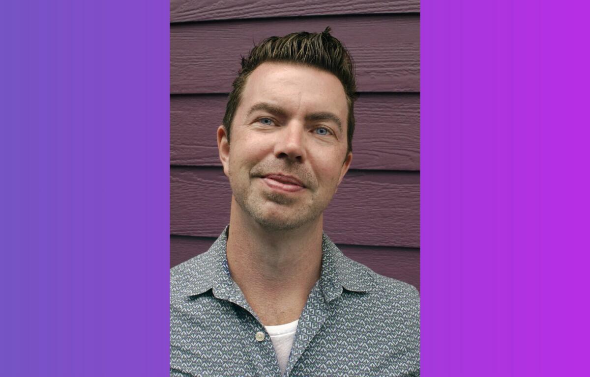 Portrait of a man with short dark hair and light stubble, wearing a patterned shirt, standing against a purple wooden wall.
