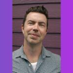 Portrait of a man with short dark hair and light stubble, wearing a patterned shirt, standing against a purple wooden wall.