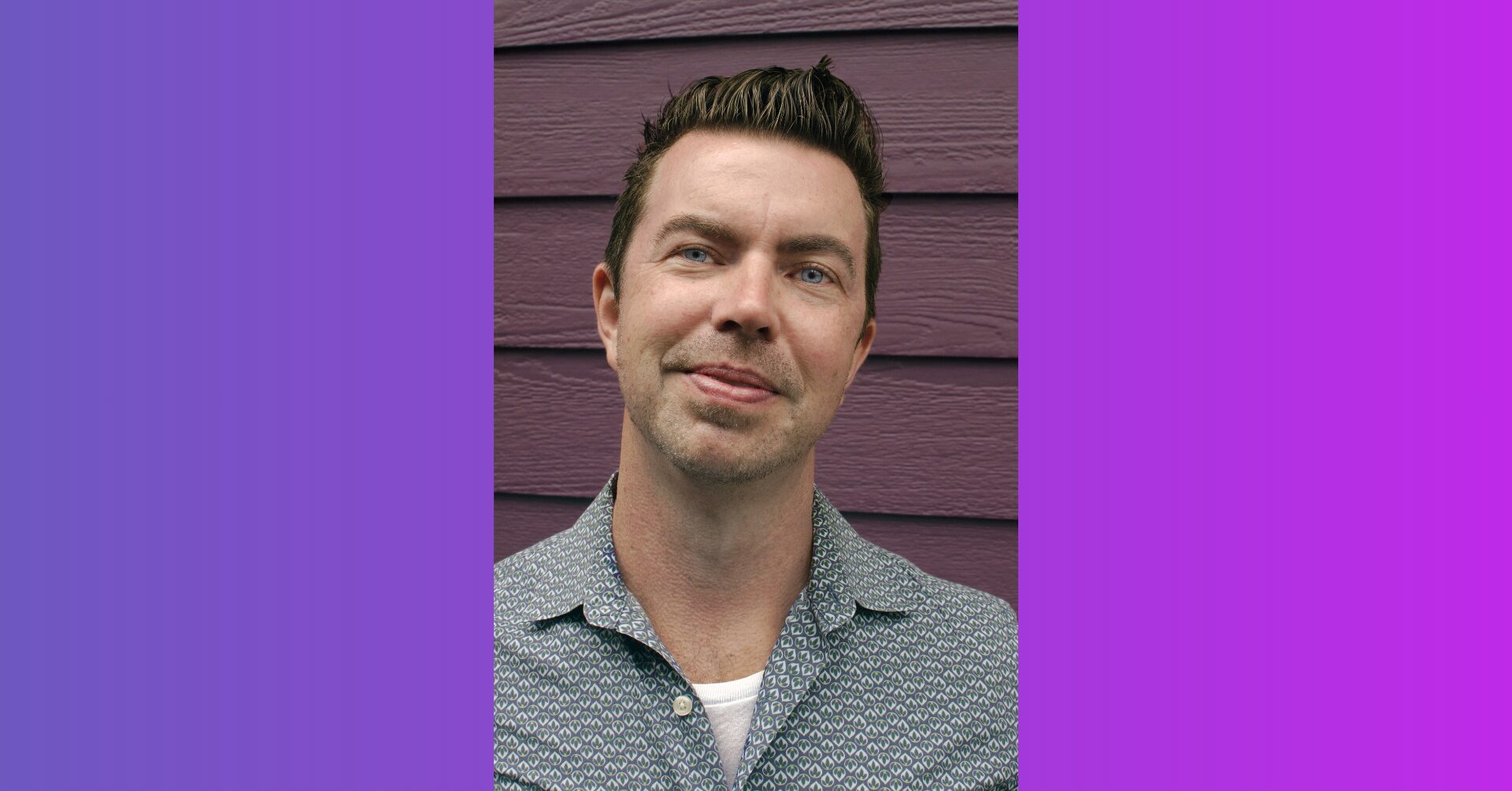 Portrait of a man with short dark hair and light stubble, wearing a patterned shirt, standing against a purple wooden wall.