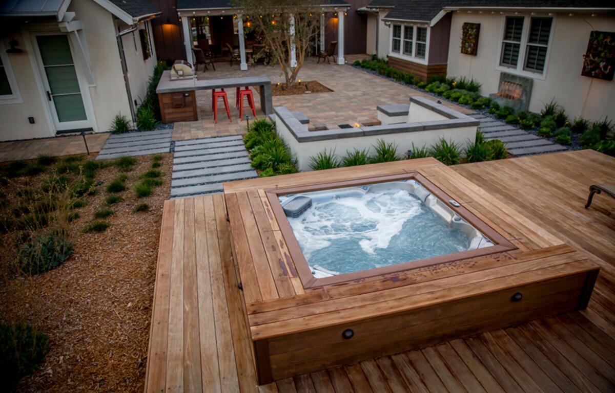 Wooden-deck backyard with a bubbling hot tub, stone patio, and surrounding greenery in a modern courtyard.