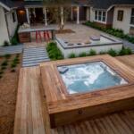 Wooden-deck backyard with a bubbling hot tub, stone patio, and surrounding greenery in a modern courtyard.