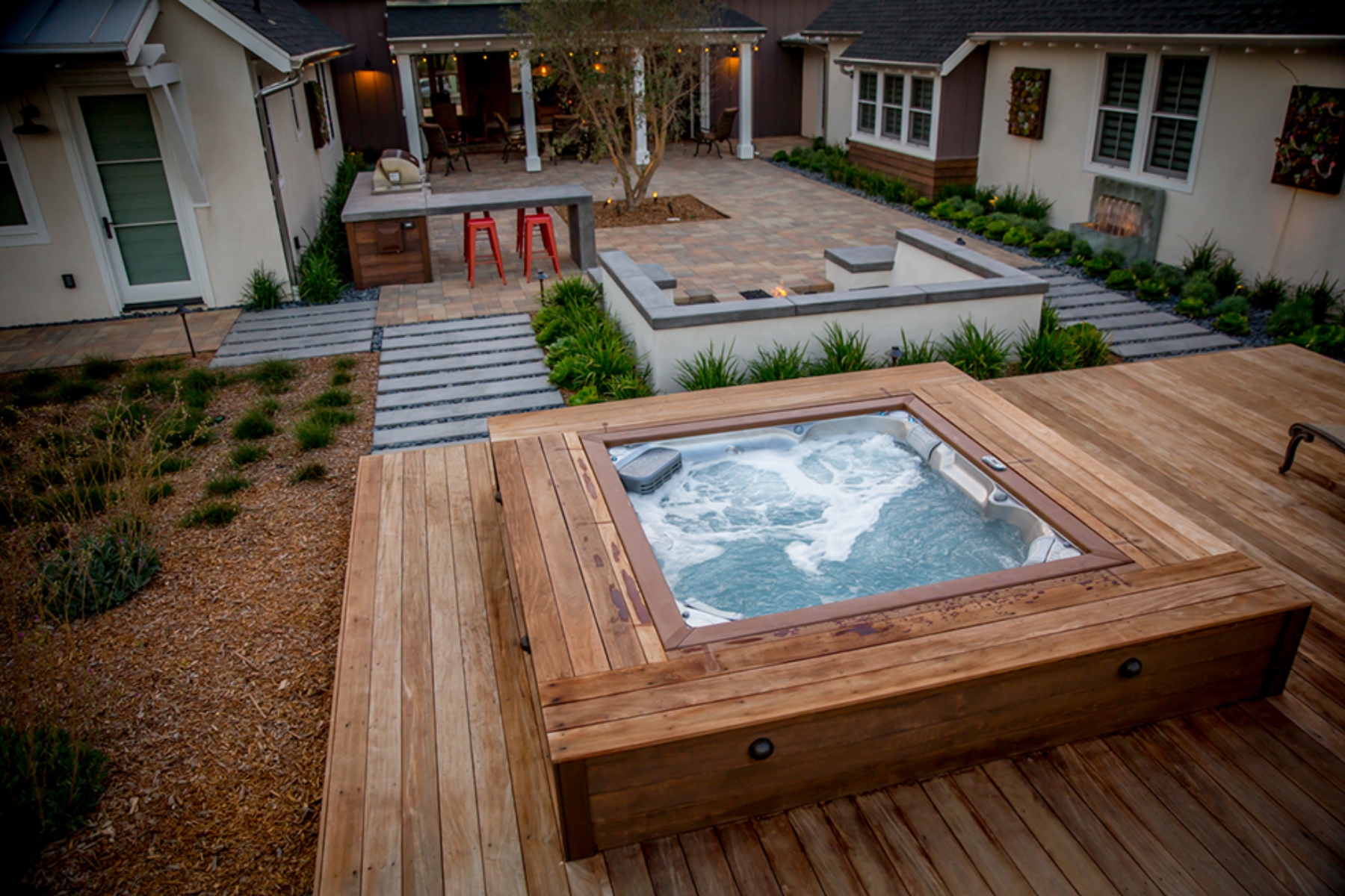 Wooden-deck backyard with a bubbling hot tub, stone patio, and surrounding greenery in a modern courtyard.