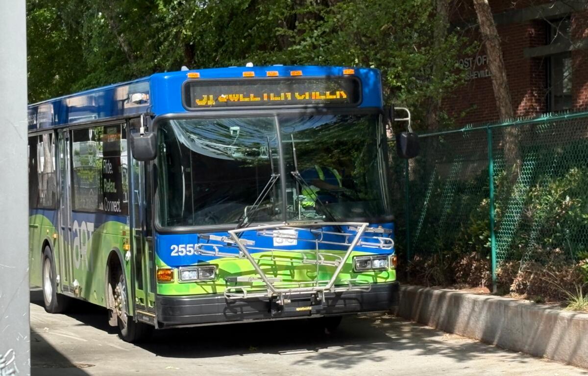 Front view of a blue and green city bus with a bike rack on the bumper and an amber destination sign above the windshield, parked on a street.