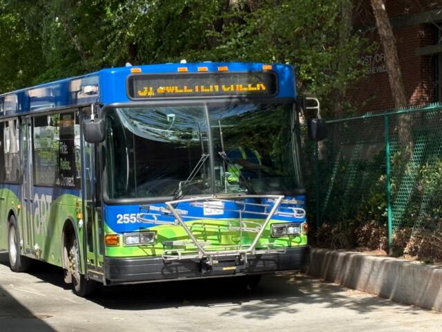 Front view of a blue and green city bus with a bike rack on the bumper and an amber destination sign above the windshield, parked on a street.